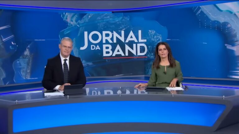 Eduardo Oinegue e Adriana Araújo, apresentadores do Jornal da Band, sentados na bancada durante telejornal com telão azul ao fundo exibindo logotipo do programa.