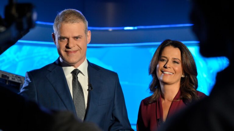 Eduardo Oinegue e Adriana Araújo sorrindo no estúdio do Jornal da Band com iluminação azul ao fundo e câmeras em primeiro plano