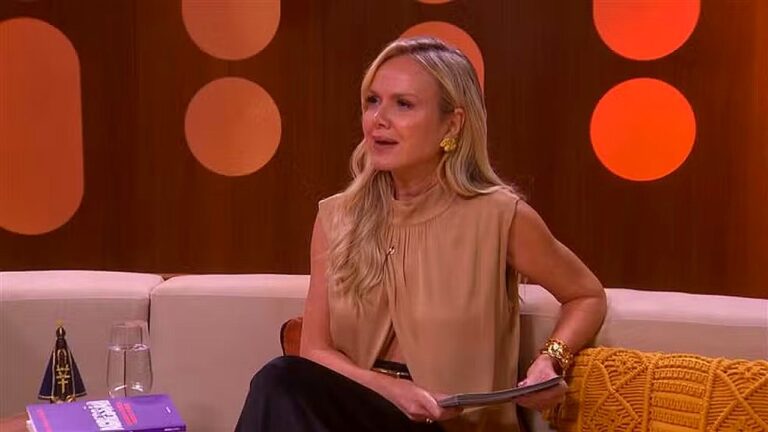 Apresentadora loira sentada em sofá fala durante programa de TV com cenário laranja ao fundo