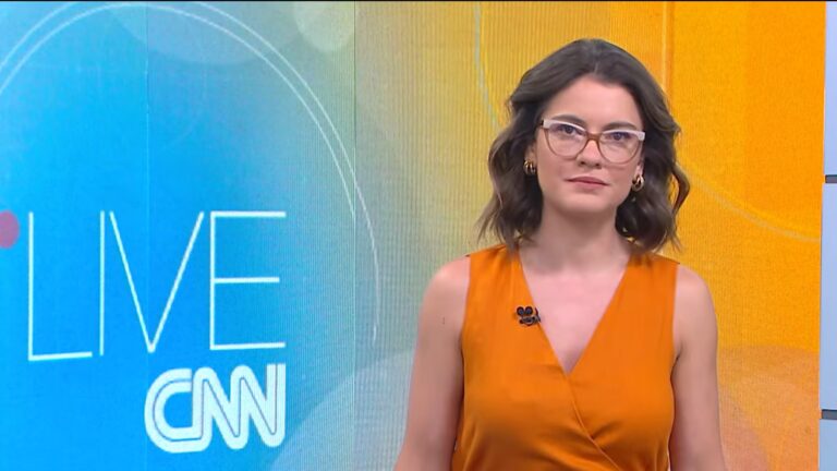 Elisa Veeck, apresentadora da CNN Brasil, usa vestido laranja e óculos durante apresentação ao vivo no estúdio do Live CNN.