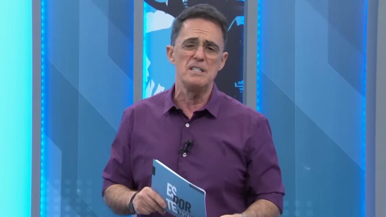 Fernando Fernandes, apresentador com camisa roxa, segura cartão do Esporte Total no estúdio da Band, com telão azul ao fundo.