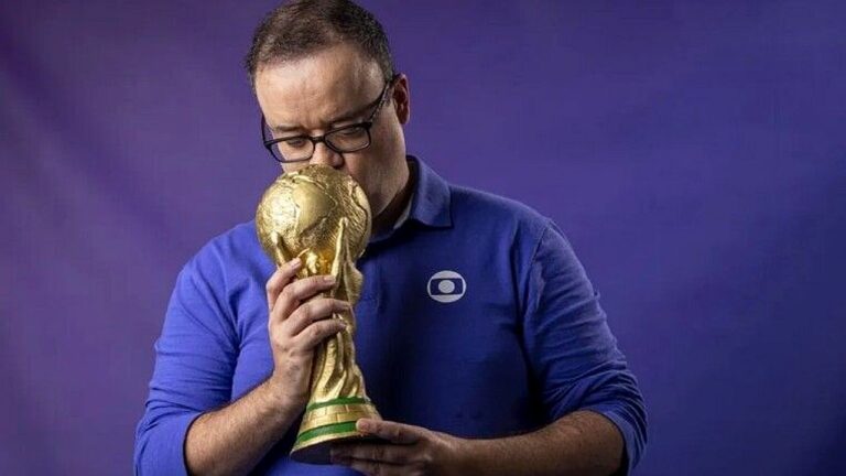 Homem de camisa da TV Globo beija réplica da taça da Copa do Mundo em fundo azul