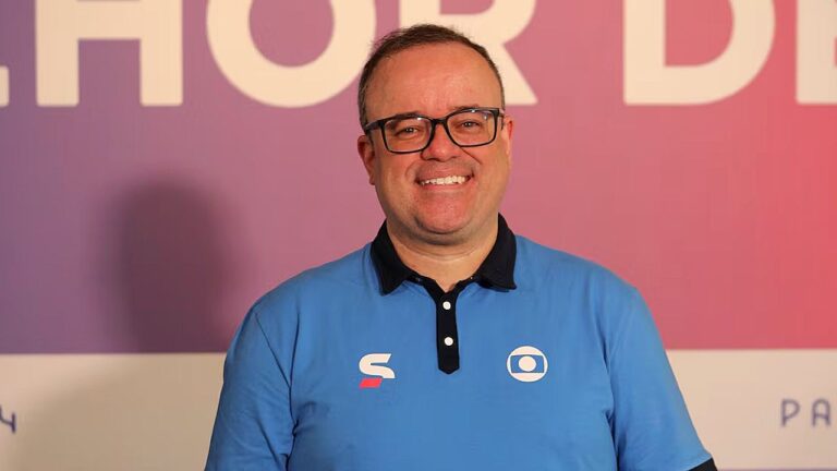 Apresentador esportivo de óculos sorri vestindo camisa azul com logotipo da Globo em fundo de evento