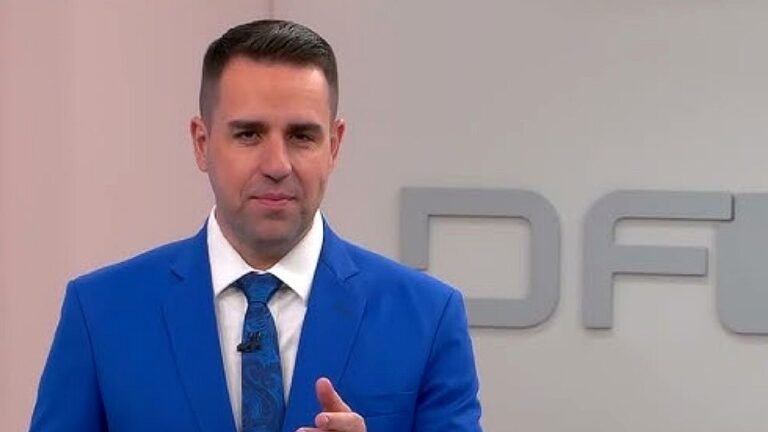 Apresentador de terno azul fala em estúdio com fundo claro e logotipo desfocado ao fundo