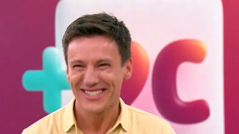 Apresentador sorri em estúdio de TV com fundo colorido e logotipo ao fundo durante programa de entretenimento