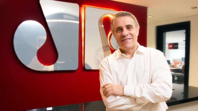 Fernando Pelegio em escritório com parede vermelha e logo da Jovem Pan em destaque ao fundo com iluminação interna