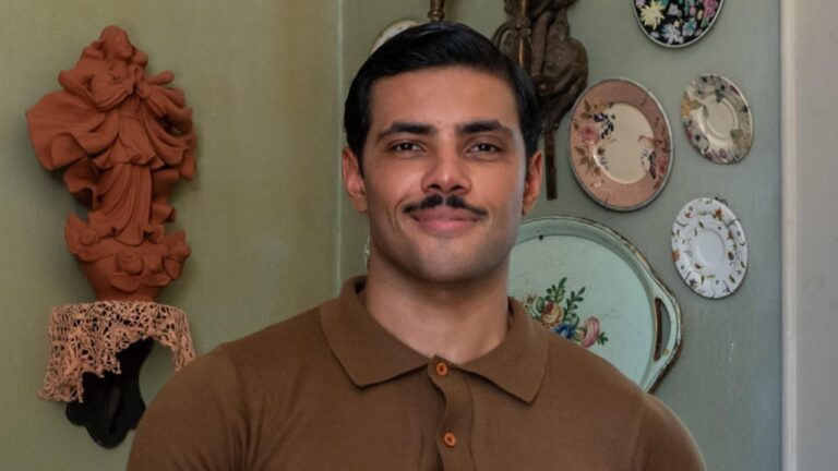 Gabriel Fuentes com bigode e camisa polo marrom posa em ambiente interno com pratos decorativos na parede ao fundo