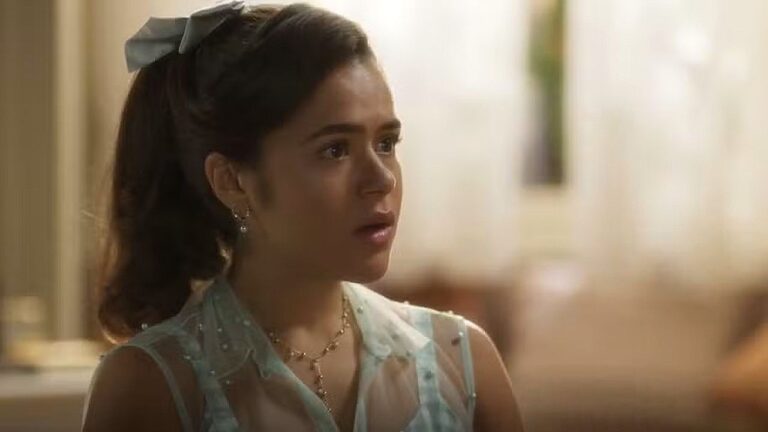 Jovem com laço no cabelo reage surpresa durante conversa em cena de novela ambientada em casa