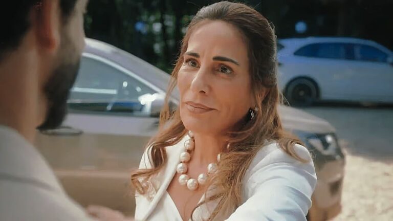 Mulher de branco com colar de pérolas conversa com homem ao lado de carro em cena de novela