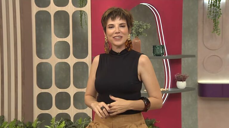 Glória Vanique sorri e fala em programa da TV Gazeta com cenário decorado e prateleiras ao fundo.