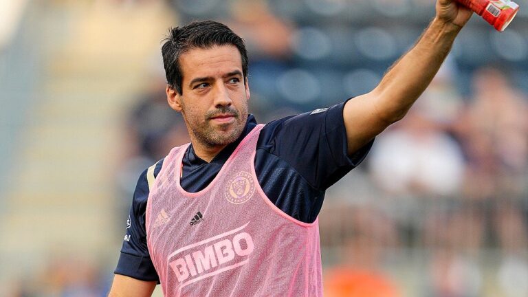 Jogador de futebol em aquecimento levanta garrafa durante treino em campo com uniforme de treino patrocinado