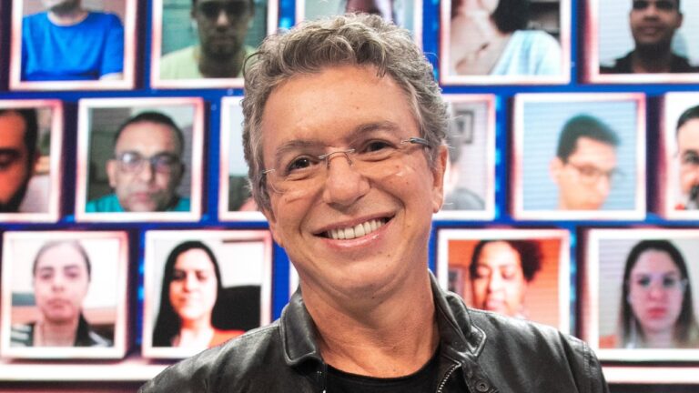 Boninho sorri diante de painel com rostos de participantes exibidos em telas durante o Domingão com Huck