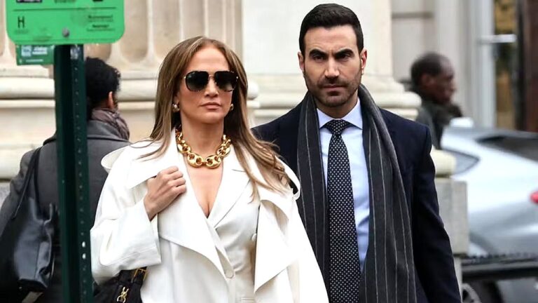 Jennifer Lopez de casaco branco e óculos escuros caminha ao lado de homem de terno em cena urbana