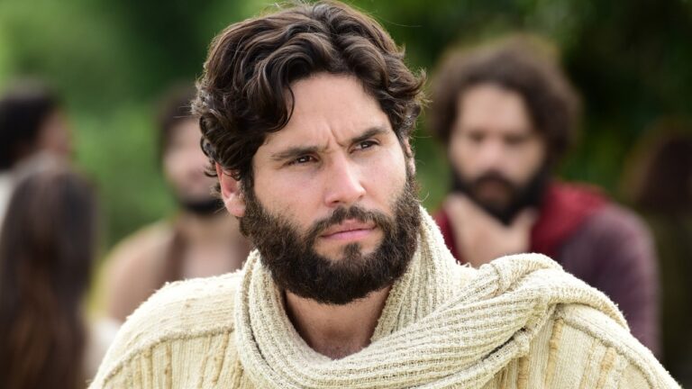 Dudu Azevedo caracterizado como Jesus com túnica e manto claro em cena bíblica ao ar livre com figurantes ao fundo