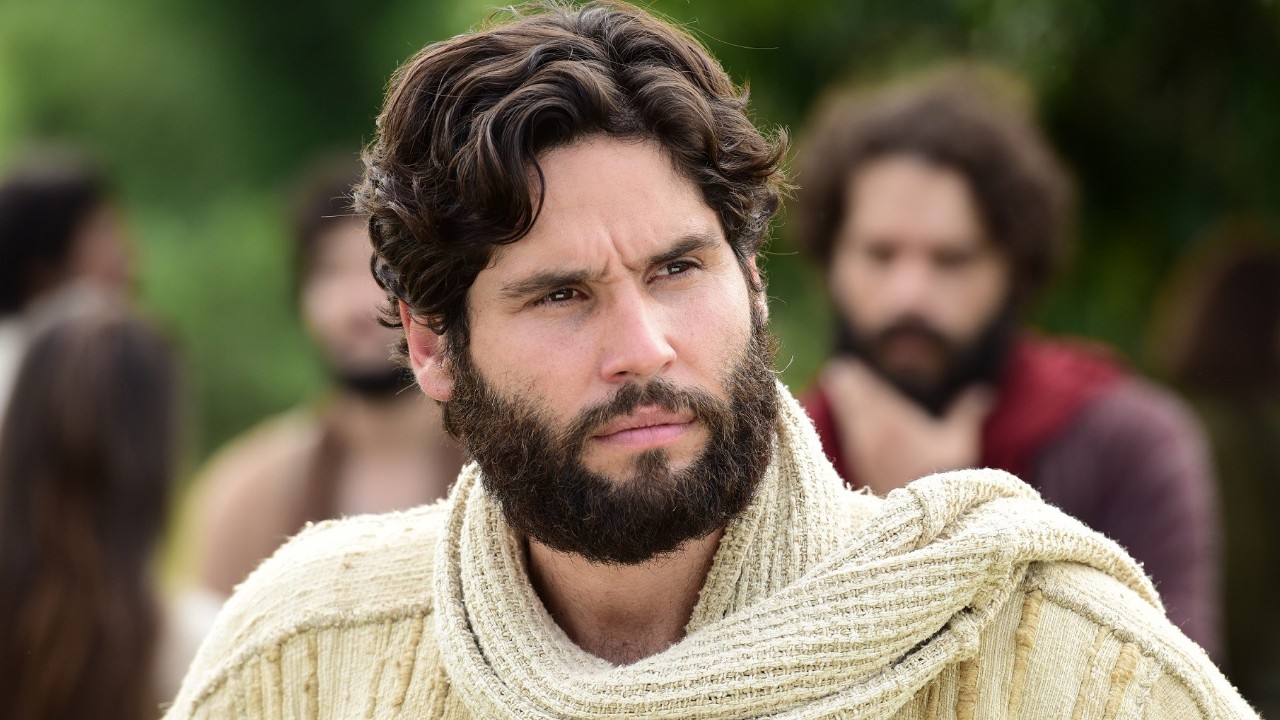 Dudu Azevedo caracterizado como Jesus com túnica e manto claro em cena bíblica ao ar livre com figurantes ao fundo