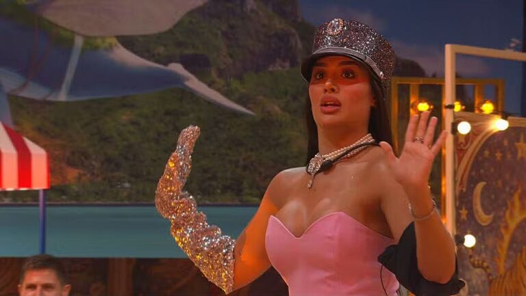 Mulher com vestido rosa e luva brilhante levanta as mãos surpresa em palco de programa de entretenimento