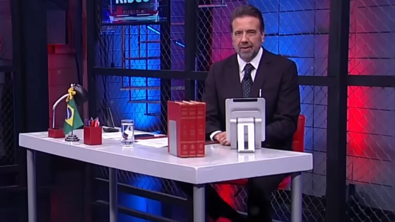 Apresentador Jorge Lordello sentado à mesa em estúdio da RedeTV! durante gravação de programa, com cenário iluminado em azul e vermelho.