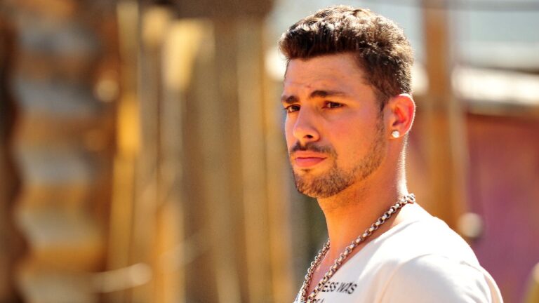 Cauã Reymond como Jorginho em Avenida Brasil com camiseta branca corrente no pescoço e olhar atento em cenário externo iluminado pelo sol