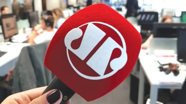 Microfone com logotipo da Jovem Pan em destaque em redação de jornalismo com profissionais e computadores ao fundo