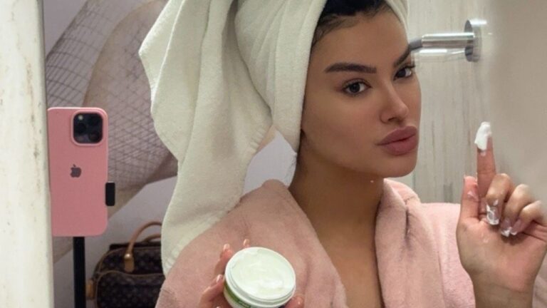 Júlia Medeiros posa em selfie no espelho com toalha na cabeça e roupão rosa segurando pote de creme e aplicando produto no dedo em banheiro