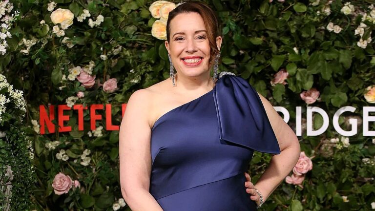 Artista posa sorrindo em evento da Netflix com vestido azul em frente a painel floral com logotipo Bridgerton