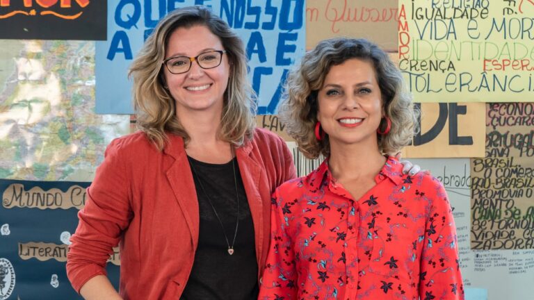 Júlia Spadaccini e Carla Faour posam sorrindo diante de mural com cartazes e palavras sobre educação e sociedade
