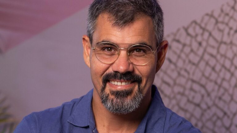 Juliano Cazarré sorri usando óculos e camisa azul em retrato com fundo neutro e iluminação suave