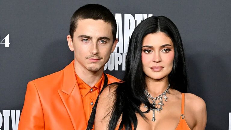 Homem e mulher posam juntos em evento com roupas laranja diante de painel com logotipo de estúdio