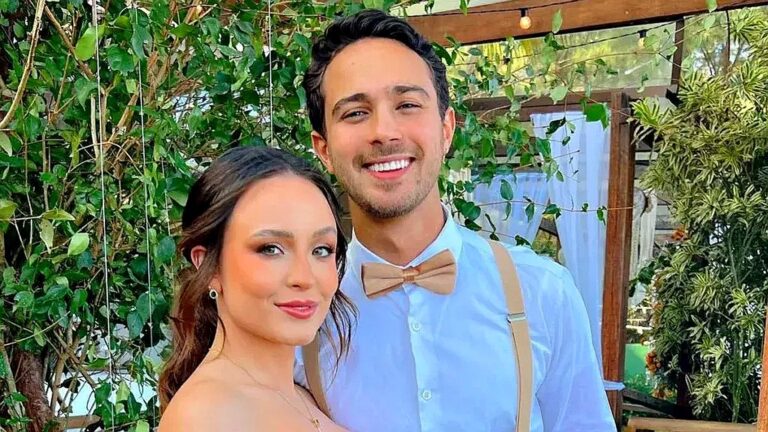 Casal sorridente posa junto em ambiente externo com plantas ao fundo em foto de evento social