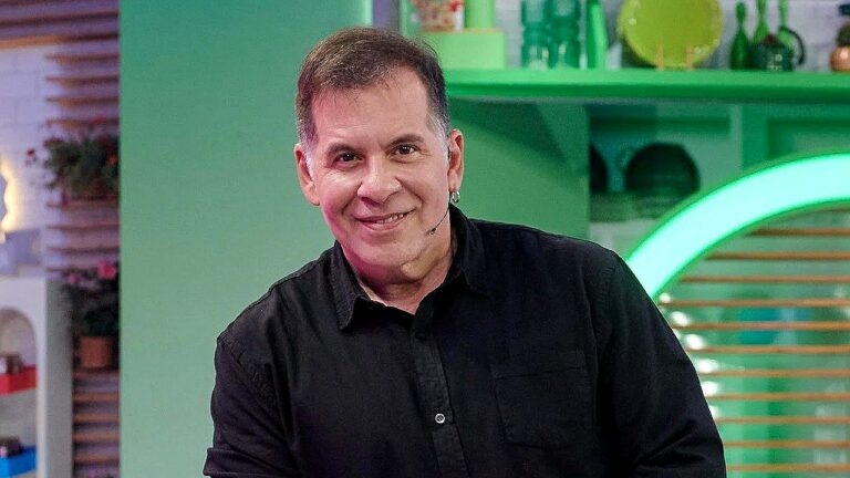 Apresentador de TV sorri em estúdio com cenário colorido e iluminação verde ao fundo