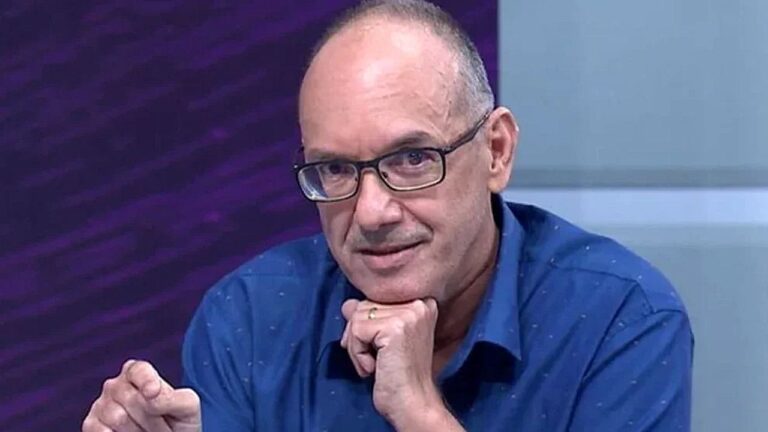 Homem de óculos apoia o queixo na mão durante participação em programa de TV em estúdio