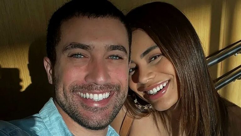 Casal sorri junto em selfie com rostos próximos em ambiente interno com iluminação quente ao fundo