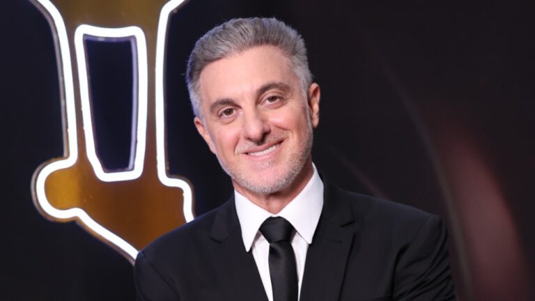 Luciano Huck sorri usando terno preto e gravata em estúdio com iluminação e elemento cenográfico ao fundo