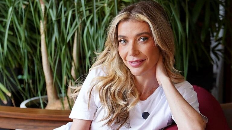 Mulher loira posa sorrindo com a mão no pescoço em ambiente com plantas ao fundo