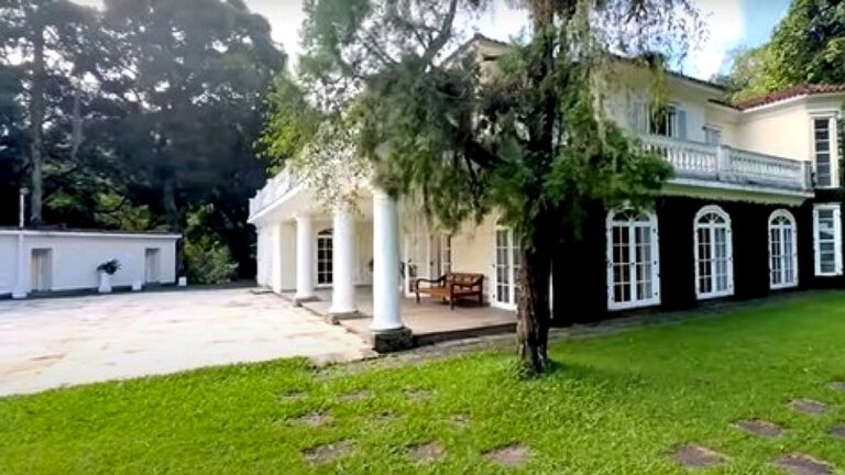 Casa grande com colunas brancas, varanda e jardim arborizado em área residencial ampla