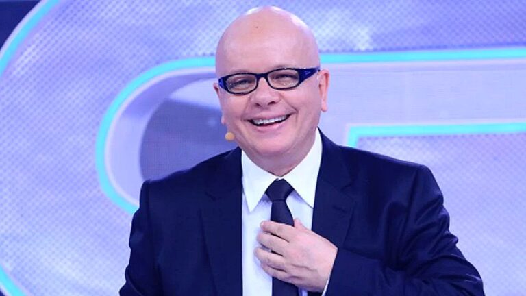 Apresentador de TV de terno e gravata sorri no estúdio enquanto ajusta a gravata durante programa ao vivo