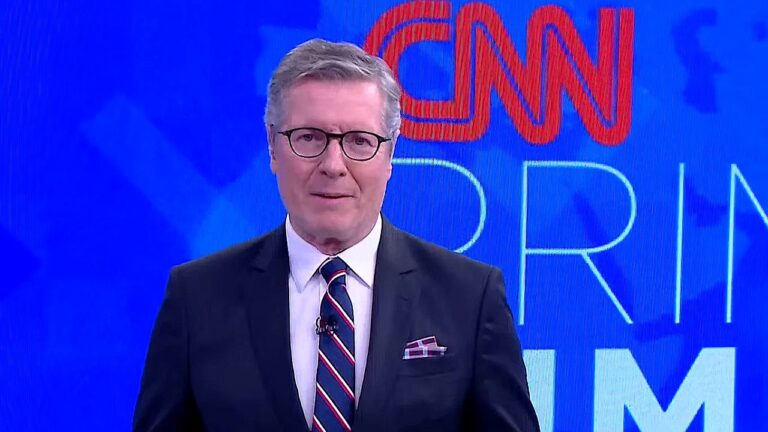 Homem de terno e óculos apresenta telejornal em estúdio com logotipo da CNN ao fundo