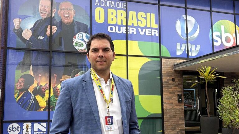 Homem com credencial posa em frente à sede do SBT com painel da Copa do Mundo e logos da emissora