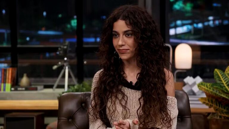 Mulher de cabelo cacheado participa de entrevista em programa noturno com cenário urbano ao fundo