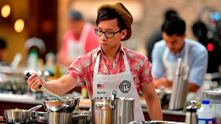 Participante com avental do MasterChef mexe panela com batedor durante prova culinária em cozinha profissional