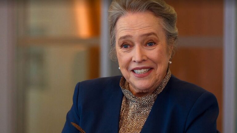 Mulher idosa sorrindo durante entrevista em ambiente interno com blazer escuro e blusa de gola alta