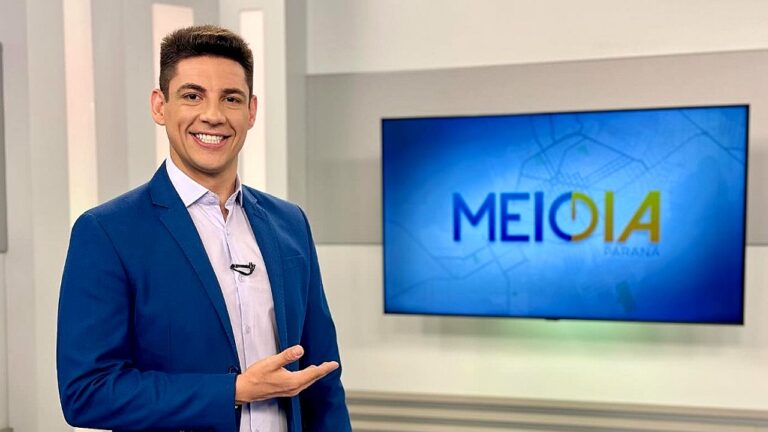 Apresentador sorri e aponta para tela com logotipo do programa Meio Dia no estúdio de TV