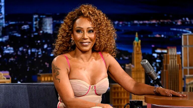 Atriz com vestido claro participa de entrevista em talk show noturno com cidade iluminada ao fundo