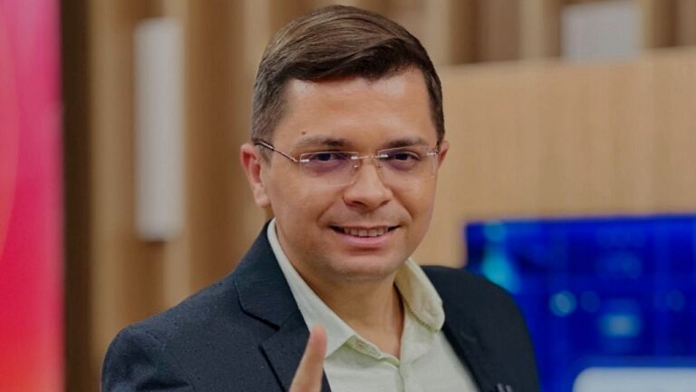 Apresentador de óculos e terno aponta para frente durante programa em estúdio com fundo desfocado