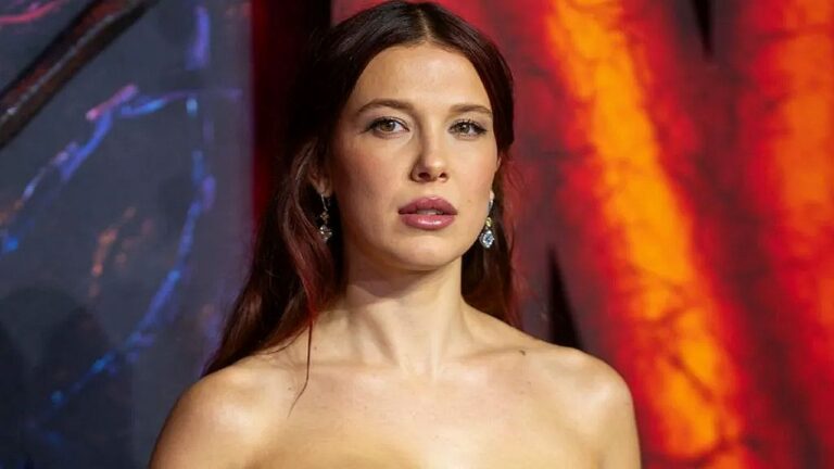 Atriz ruiva usa vestido sem alças e brincos em evento com fundo iluminado em tons de vermelho