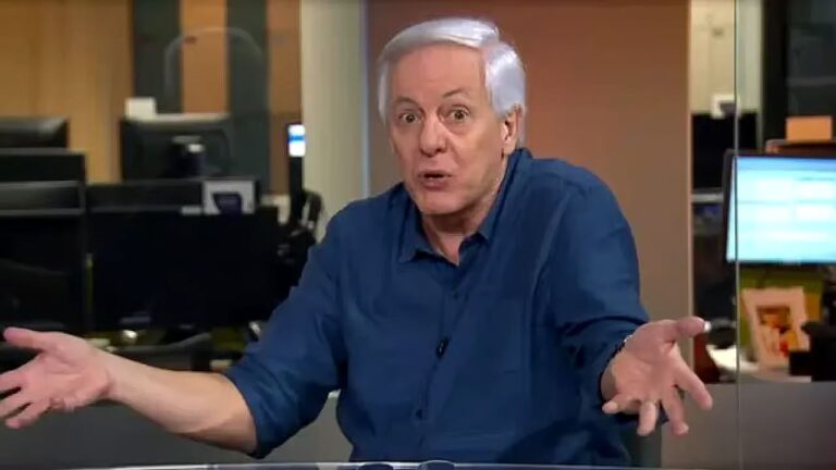 Texto Alternativo disse: Apresentador de camisa azul gesticula com as mãos abertas durante fala em estúdio de jornalismo com computadores ao fundo