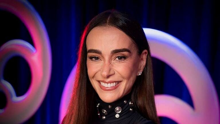 Atriz sorrindo em evento com fundo iluminado e logotipo estilizado ao fundo em cenário de premiação
