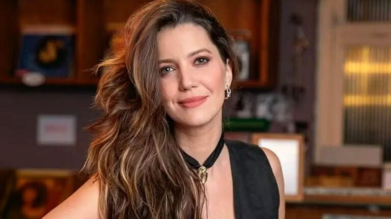 Mulher de cabelo longo ondulado usa blusa preta e colar no pescoço em ambiente interno com prateleiras ao fundo