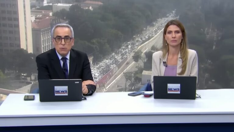 Nelson Gomes e Patricia Rocha apresentam telejornal da BandNews com laptops sobre bancada e trânsito intenso ao fundo