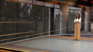 Apresentadora fica de braços cruzados em estúdio virtual do Bom Dia Brasil com telões curvos e iluminação em tons laranja